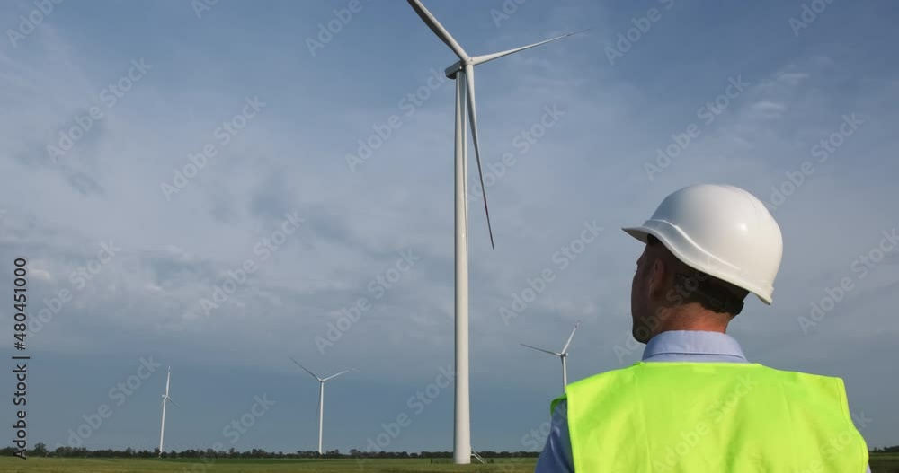 A man in a white protective helmet goes to the wind turbine, raises an electronic tablet in front of him, the worker maintains the operation of the wind generator. A man in a white protective helmet goes to the wind turbine, raises an electronic tablet in front of him, the worker maintains the operation of the wind generator.