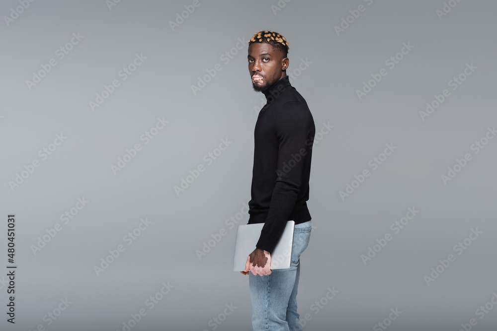 trendy african american man with vitiligo standing with laptop and looking at camera isolated on grey. trendy african american man with vitiligo standing with laptop and looking at camera isolated on grey.