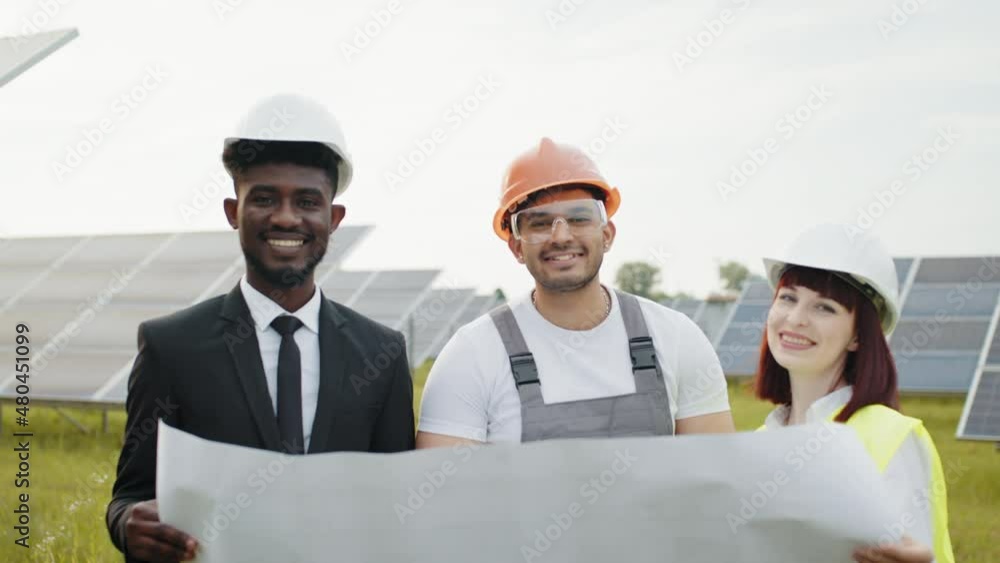 Engineers at solar farm. Two engineers and one technician examining drawings of solar farm while standing together outdoors. Male and female partners in helmets working on modern business project Engineers at solar farm. Two engineers and one technician examining drawings of solar farm while standing together outdoors. Male and female partners in helmets working on modern business project