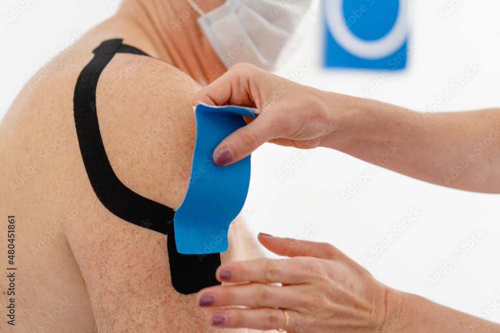 close-up of a therapist putting Elastic Therapeutic Kinesiology Tape on an elderly man's shoulder to recover from tendonitis close-up of a therapist putting Elastic Therapeutic Kinesiology Tape on an elderly man's shoulder to recover from tendonitis
