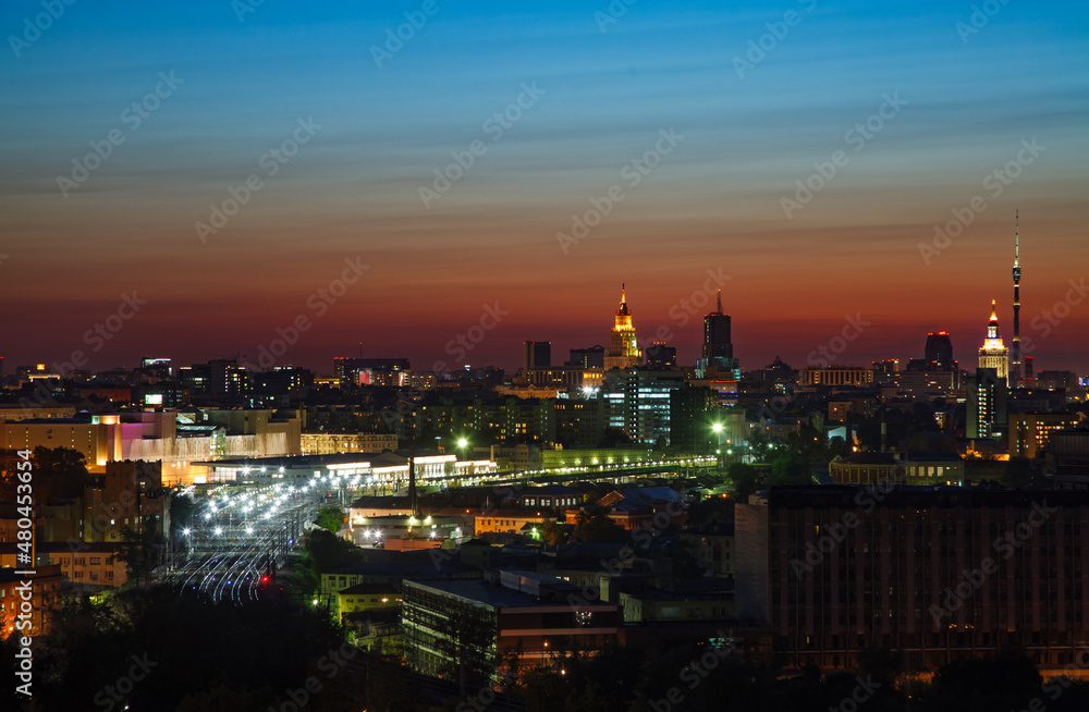 Red and blue saturated sunset with light clouds in the city. Silhouette of town and lights of railway station and railroad. Moscow, Russia.