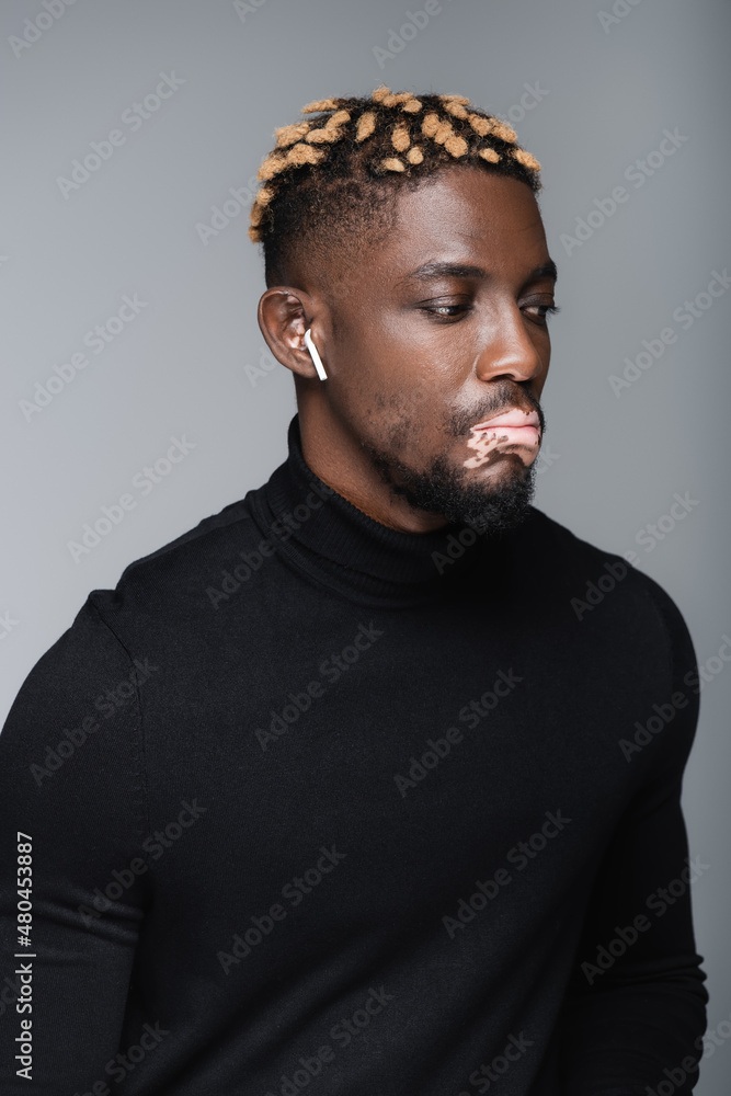 young african american man with vitiligo and trendy hairstyle listening music in earphone isolated on grey. young african american man with vitiligo and trendy hairstyle listening music in earphone isolated on grey.
