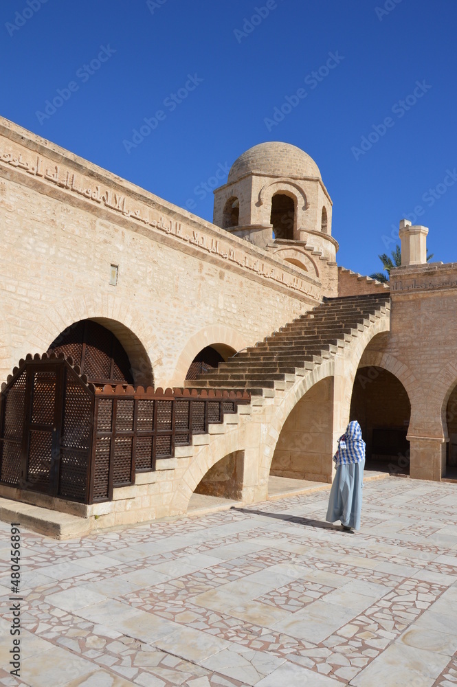 Muslim woman with covered face in the Great Mosque in Sousse, Tunisia, Africa. Blue sky, ancient stone arces, stairway and corner tower	