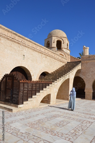 Muslim woman with covered face in the Great Mosque in Sousse, Tunisia, Africa. Blue sky, ancient stone arces, stairway and corner tower	