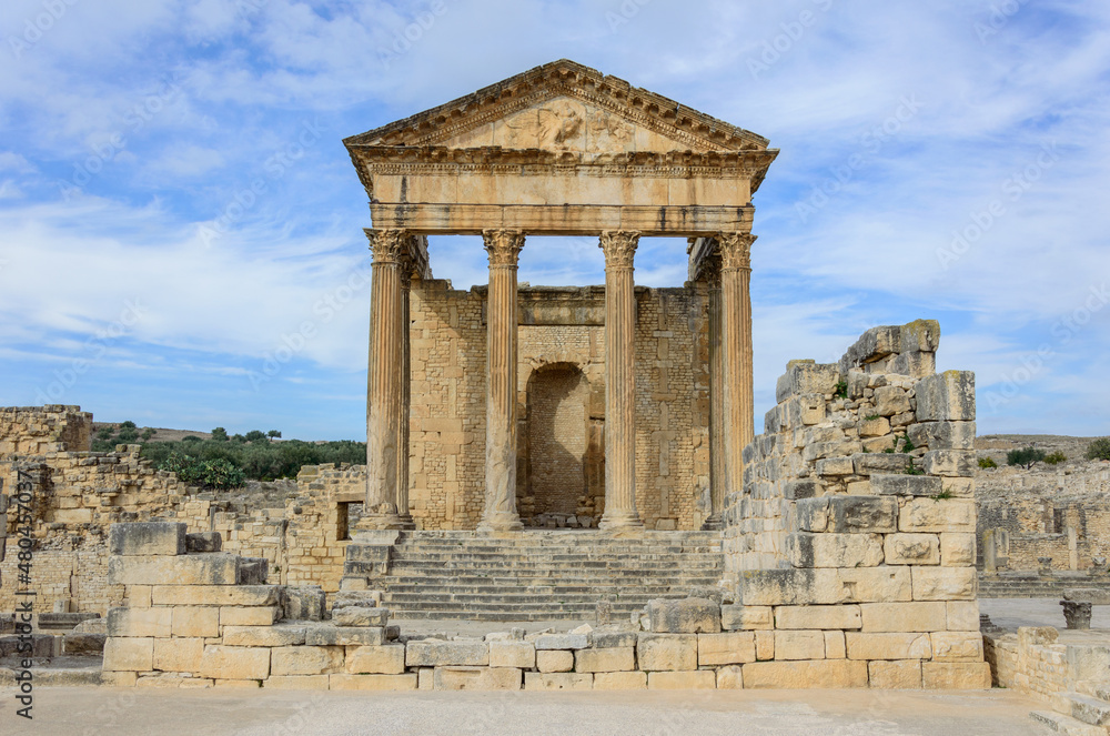 Temple of Jupiter, forum and ancient roman ruins of Dougga in Tunisia, Africa in the sunny afternoon. Blue sky with clouds, old yellow, grey and brown stone walls and columns