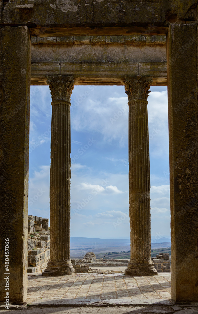 Fototapeta premium View out of the temple of Jupiter in the ancient roman ruined city of Dougga in Tunisia, Africa in the sunny afternoon. Blue sky with clouds, old yellow, grey and brown stone walls and columns