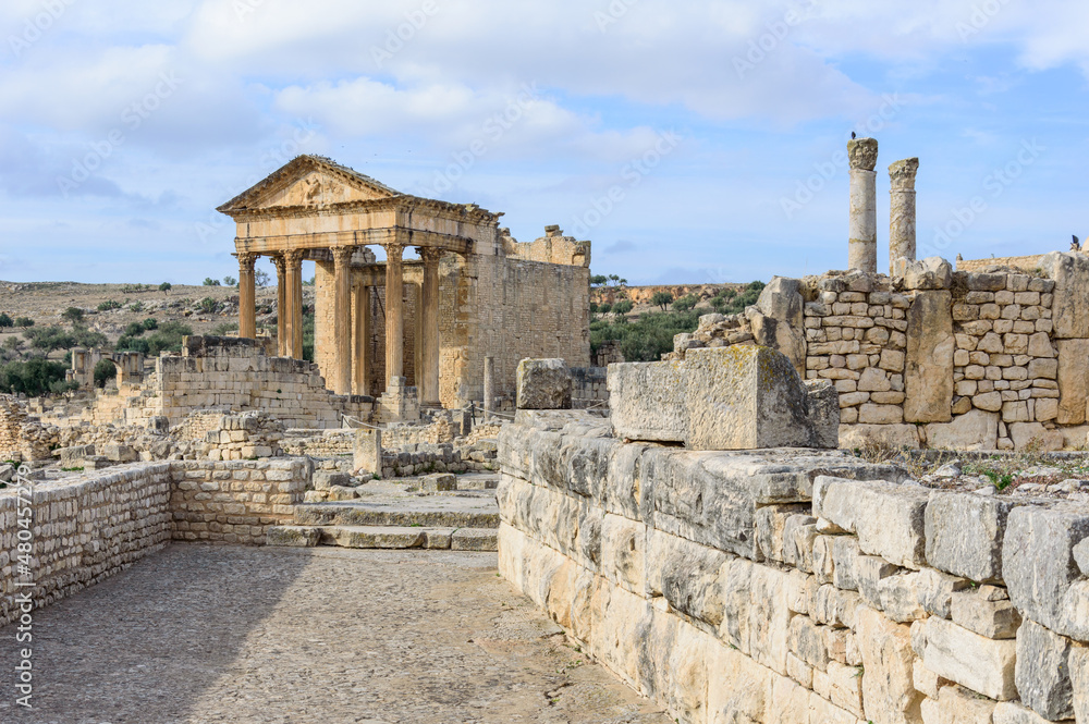 Temple of Jupiter, forum and ancient roman ruins of Dougga in Tunisia, Africa in the sunny afternoon. Blue sky with clouds, old yellow, grey and brown stone walls and columns, paved street