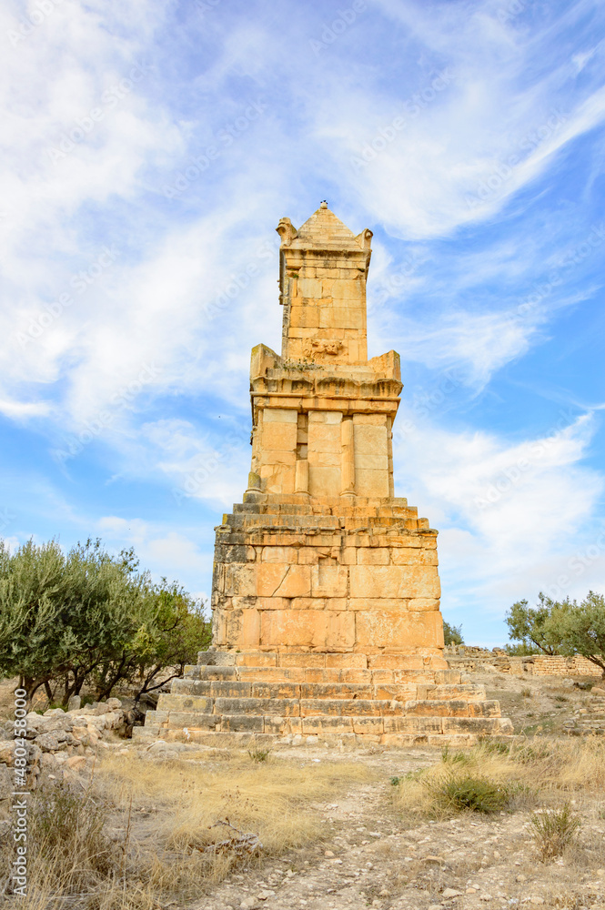 tombstone of nobleman in Thugga archeological site, Tunisia, Africa. blue sky with clouds, yellow sandstone tower, green trees, dry grass tombstone of nobleman in Thugga archeological site, Tunisia, Africa. blue sky with clouds, yellow sandstone tower, green trees, dry grass