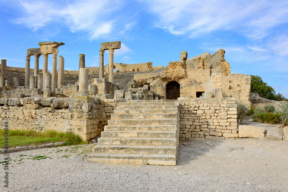Ruins of the ancient Roman theatre in Dougga, Tunisia, Africa.  Blue sky with clouds, old yellow, grey and brown stone walls, arches and columns with capitels