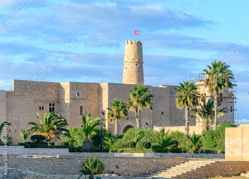 Ancient arabic ribat fortress walls and tower in Monastir, Tunisia, Africa. blue sky with clouds, yellow stones, green palm trees, sunlight on the walls, red tunisian in the sky above the tower photo