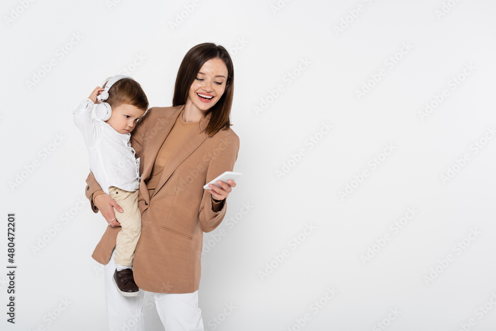 happy woman in beige blazer holding smartphone and toddler boy in headphones isolated on grey. happy woman in beige blazer holding smartphone and toddler boy in headphones isolated on grey.
