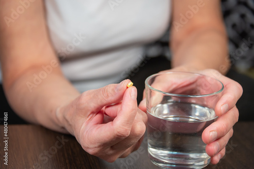 The woman holds a small pill and a glass of water in her hand. The woman is drinking medicine.
