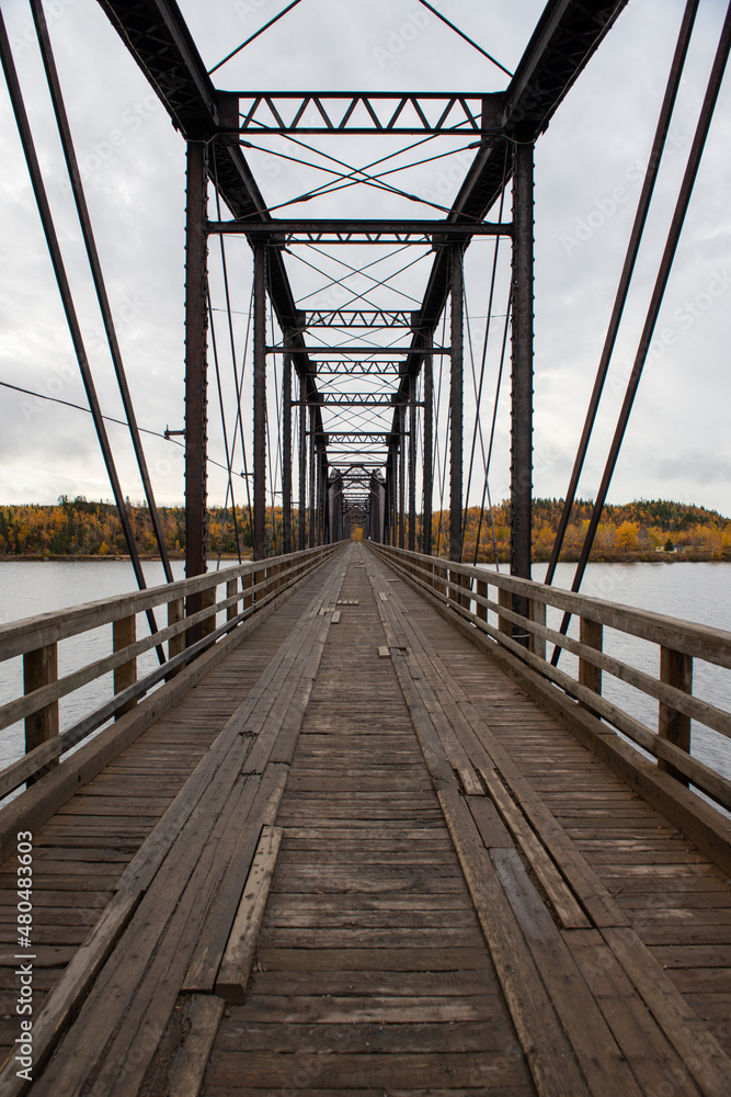Obraz premium Steel trestle bridge with wooden deck over a large river in Newfoundland. The bridge is for foot traffic and ATV usage. The sky is clear blue and land and houses can be seen in the background.