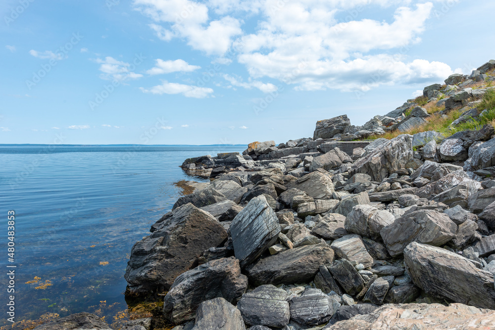 The smooth crystal clear ocean water under blue sky with clouds. The shoreline is covered in large boulders, rocks, and stones moving up the grassy berm. There's land on the horizon. The smooth crystal clear ocean water under blue sky with clouds. The shoreline is covered in large boulders, rocks, and stones moving up the grassy berm. There's land on the horizon.
