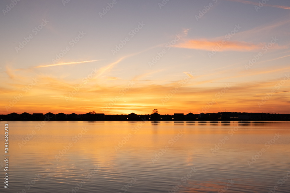 A bright orange sunset over the ocean with wispy clouds. The sky has some dark clouds and the sun is just going down over the horizon. There are water ripples in the foreground of the photo. A bright orange sunset over the ocean with wispy clouds. The sky has some dark clouds and the sun is just going down over the horizon. There are water ripples in the foreground of the photo.