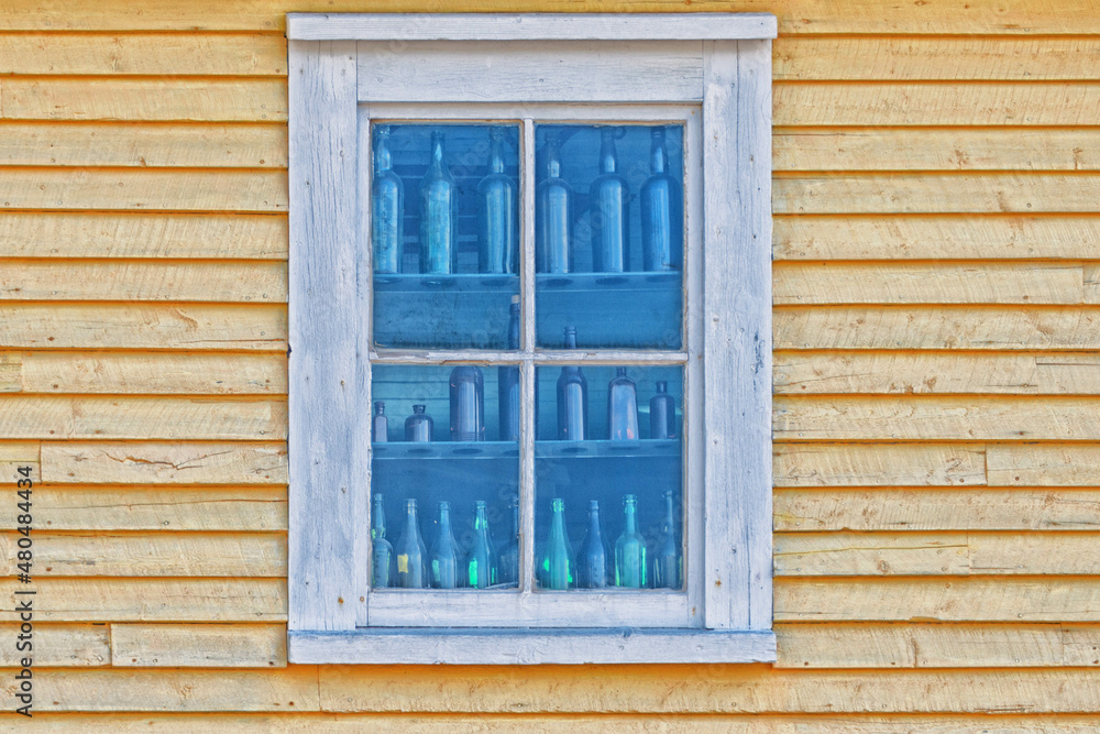 The top corner of a yellow wooden building. There's a double hung window with white trim. The edge of the building has white trim. The window has four colorful shelves lined with vintage bottles.  