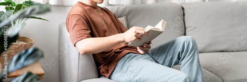 Technology Concept The male with his glasses sitting on the grey sofa, leaning his back on it, and reading a book