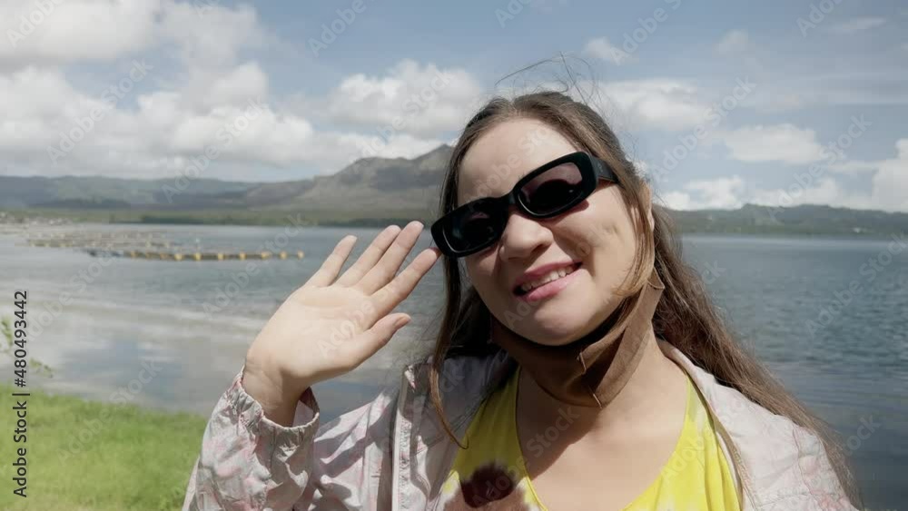 An Asian girl in fashionable sunglasses looks into camera and waves against background of lake and large volcano with blue sky and clouds. Cheerful traveler girl on background of mountain in daytime. An Asian girl in fashionable sunglasses looks into camera and waves against background of lake and large volcano with blue sky and clouds. Cheerful traveler girl on background of mountain in daytime.
