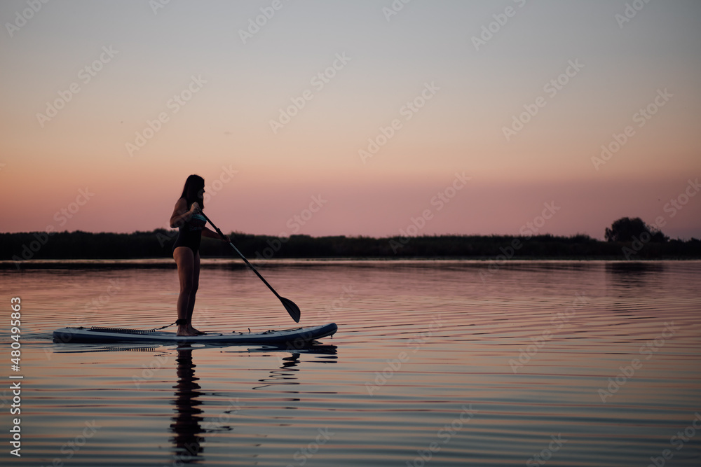 Female middle aged person with oar in hands rowing straight on evening lake with pink sky in background in swimsuit in summer. Active lifestyle for older people.