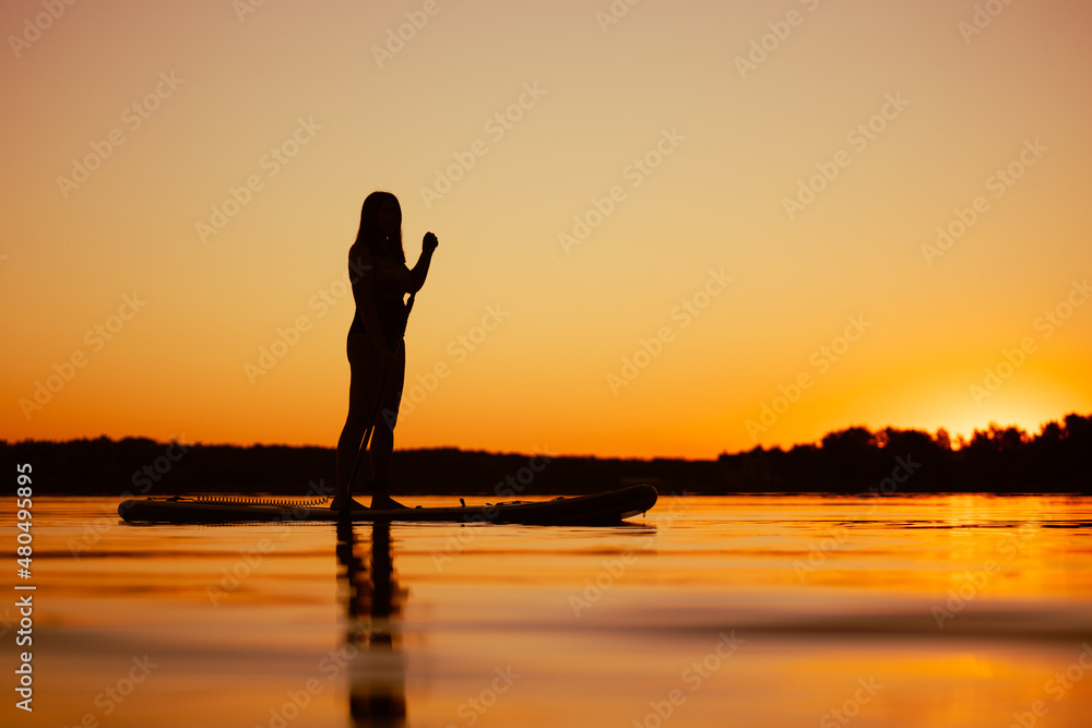 Woman shot from low angle on sup board with paddle in hands looking at amazing sunset covering water surface with orange color in evening. Active lifestyle.
