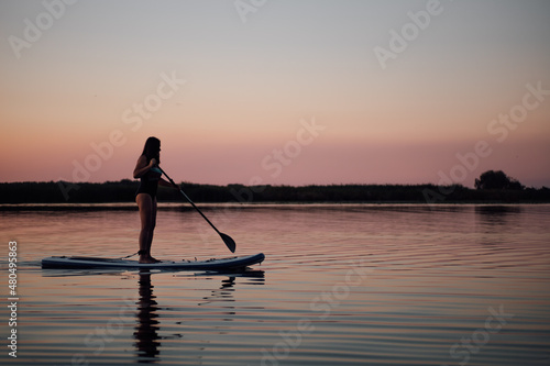 Female middle aged person with oar in hands rowing straight on evening lake with pink sky in background in swimsuit in summer. Active lifestyle for older people.