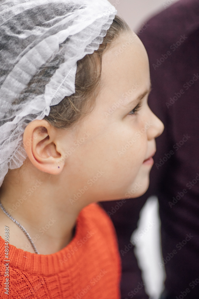 Fototapeta premium Close-up portrait in profile of face of little girl in disposable medical cap waiting for medical procedures in hospital