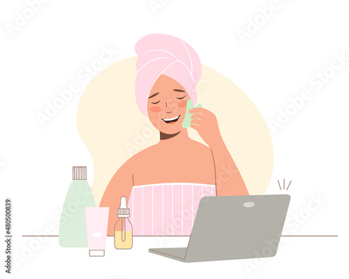 Concept of online beauty treatments. View online classes on laptop. Young woman with towel on her head takes care of her face. She uses jade gouache scraper for massage.