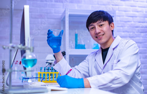 Portrait studio shot of Asian professional male scientist in white lab coat rubber gloves sitting smiling look at camera holding showing sample test tube in hands in hospital laboratory working room