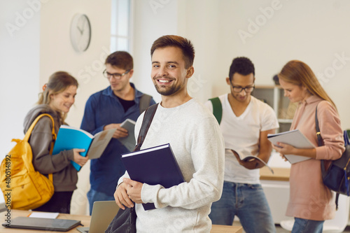 Portrait of cheerful male university student in classroom. Handsome young man with happy face expression holding textbook and smiling at camera while his classmates are reading books in background