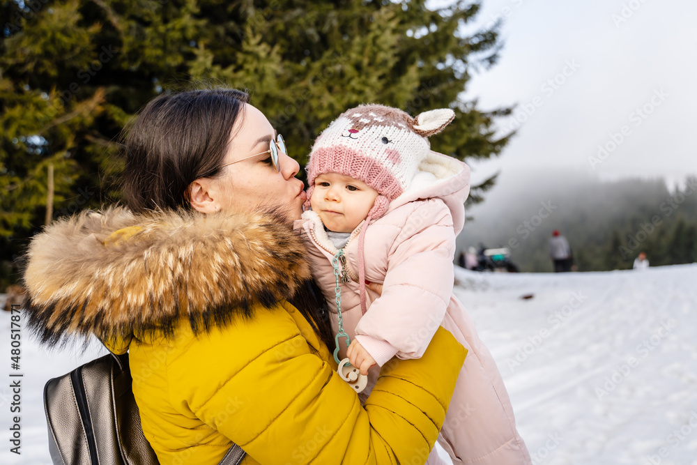 Obraz premium One caucasian woman and baby mother holding her daughter kissing her while standing outdoor in winter day with snow on mountain range parenthood family happiness concept copy space