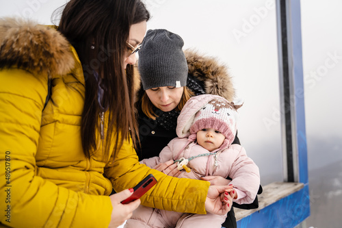 Two caucasian women standing with the baby mother and aunt holding small caucasian child while sitting tourist in winter day on mountain range with snow in nature family parenthood growing up concept