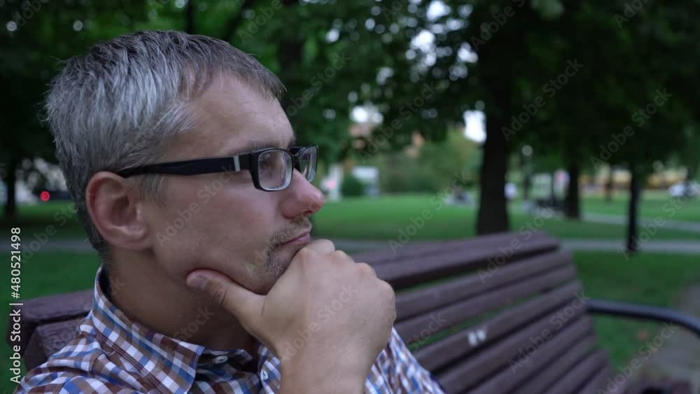 Adult grey-haired man with three-day stubble in glasses and short-sleeved shirt checks time sitting on bench and enjoying weather in park Adult grey-haired man with three-day stubble in glasses and short-sleeved shirt checks time sitting on bench and enjoying weather in park