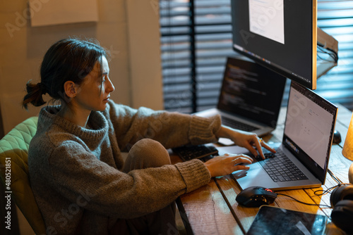 Young focused woman feeling overworked and tired, working on laptop and desktop computers, sitting at home office during the nighttime. Female programmer writing code