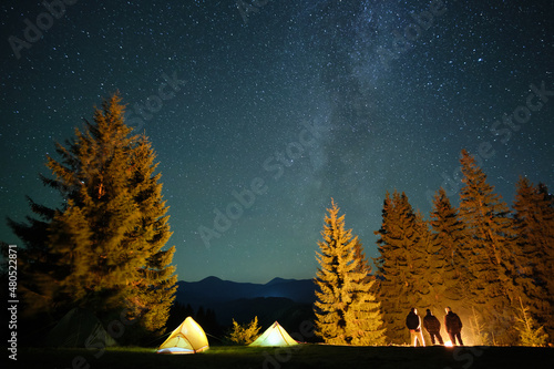 Hikers resting besides bright glowing bonfire near illuminated tourist tents on camping site in dark mountain woods under night sky with sparkling stars. Active lifestyle and outdoor living concept