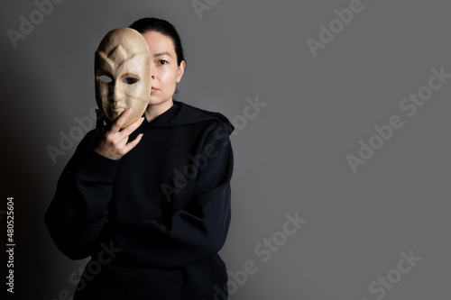 Tableau sur toile Hiding behind a mask, a young woman in a dark hoodie hides her face with a mask, the concept of self-doubt