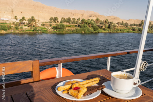 A saucer with cupcakes, pastries and a cup of coffee stands on a wooden table on the deck of a cruise liner. Behind the railing you can see the river, green vegetation and sand dunes against the sky.