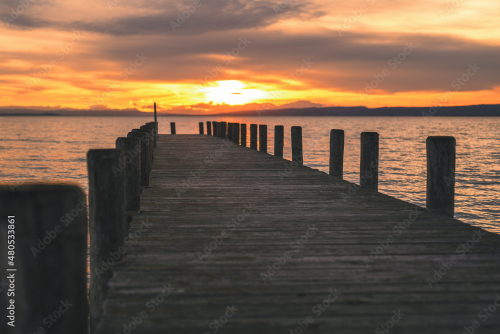 Wooden footbridge with pilings at sunset on Lake Neusiedl. The orange sun reflects in the waves of the water. The cloudy sky makes the photo very atmospheric.