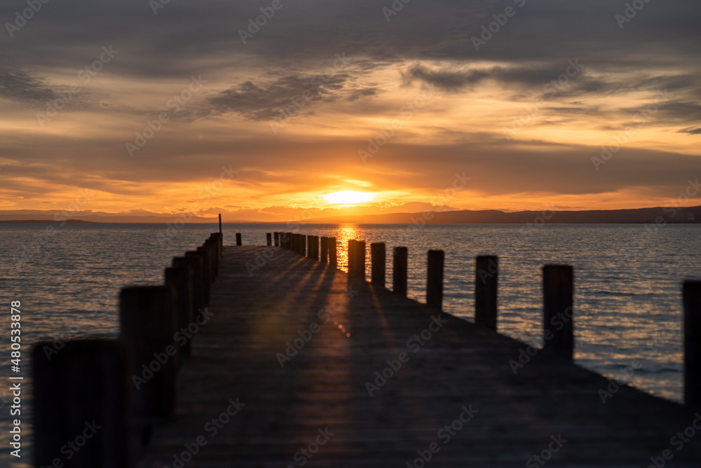 Atmospheric photo of the sunset at Lake Neusiedl with a wooden pier in the foreground. The wooden poles cast a shadow on the jetty. The orange cloudy sky with the turquoise wavy water
