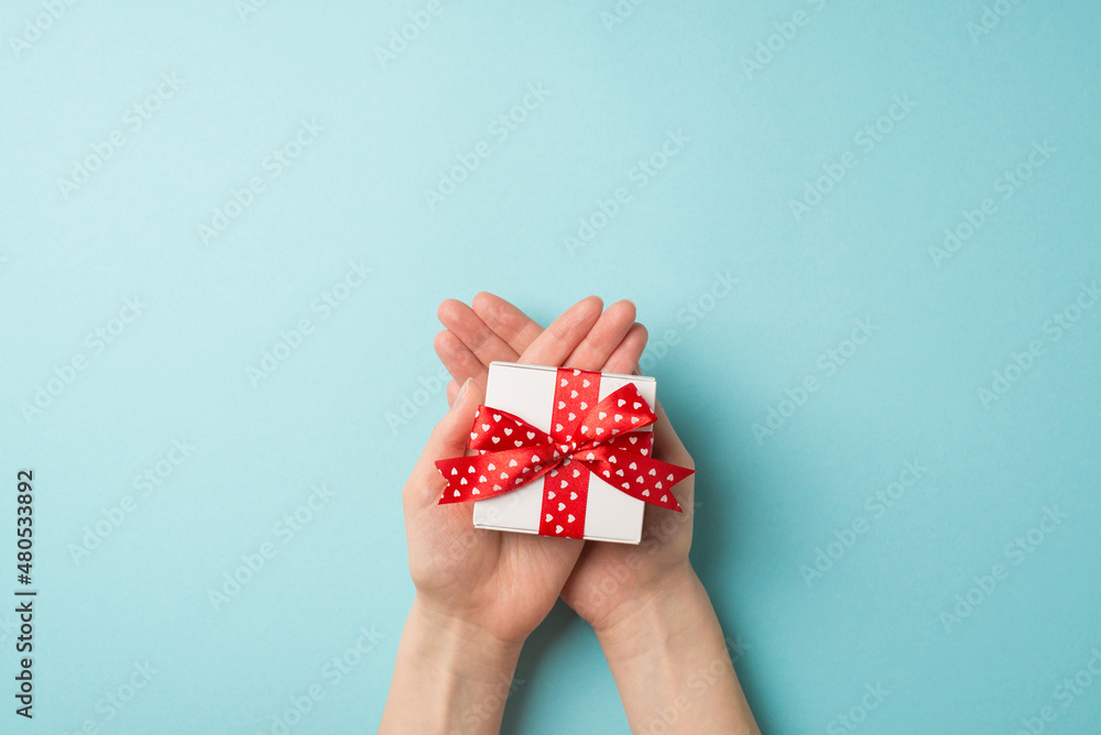 First person top view photo of valentine's day decorations young person's hands holding small white giftbox with red ribbon bow on crossed palms on isolated pastel blue background with copyspace First person top view photo of valentine's day decorations young person's hands holding small white giftbox with red ribbon bow on crossed palms on isolated pastel blue background with copyspace