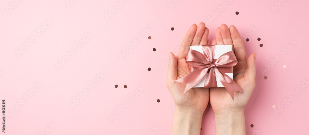 First person top view photo of valentine's day decorations woman's hands holding small white giftbox with pink ribbon bow on palms and confetti on isolated pastel pink background with empty space
