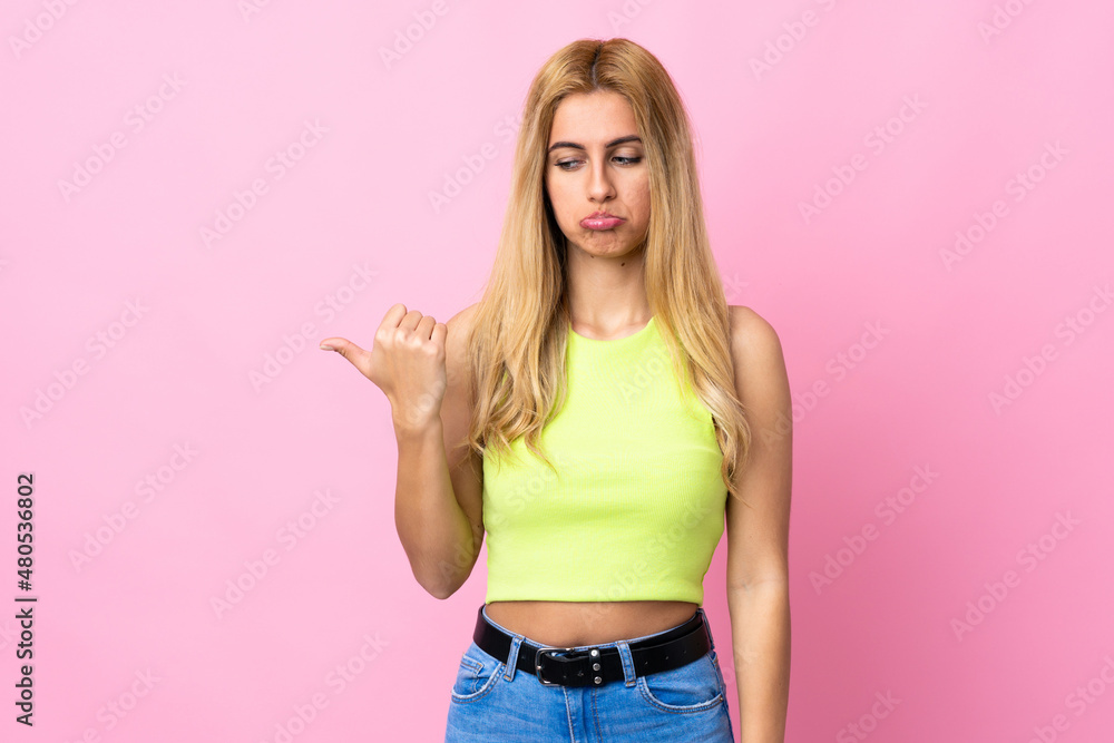 Young Uruguayan blonde woman over isolated pink background unhappy and pointing to the side Young Uruguayan blonde woman over isolated pink background unhappy and pointing to the side