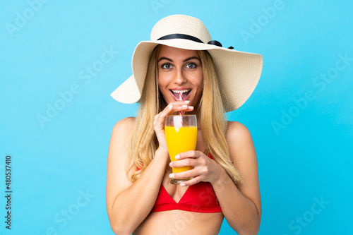 Young Uruguayan blonde woman over isolated blue background in swimsuit and holding a cocktail