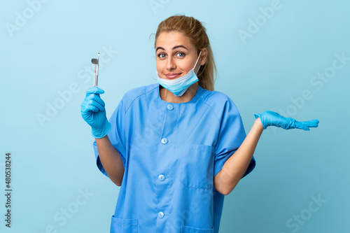 Young woman dentist holding tools over isolated blue background having doubts with confuse face expression