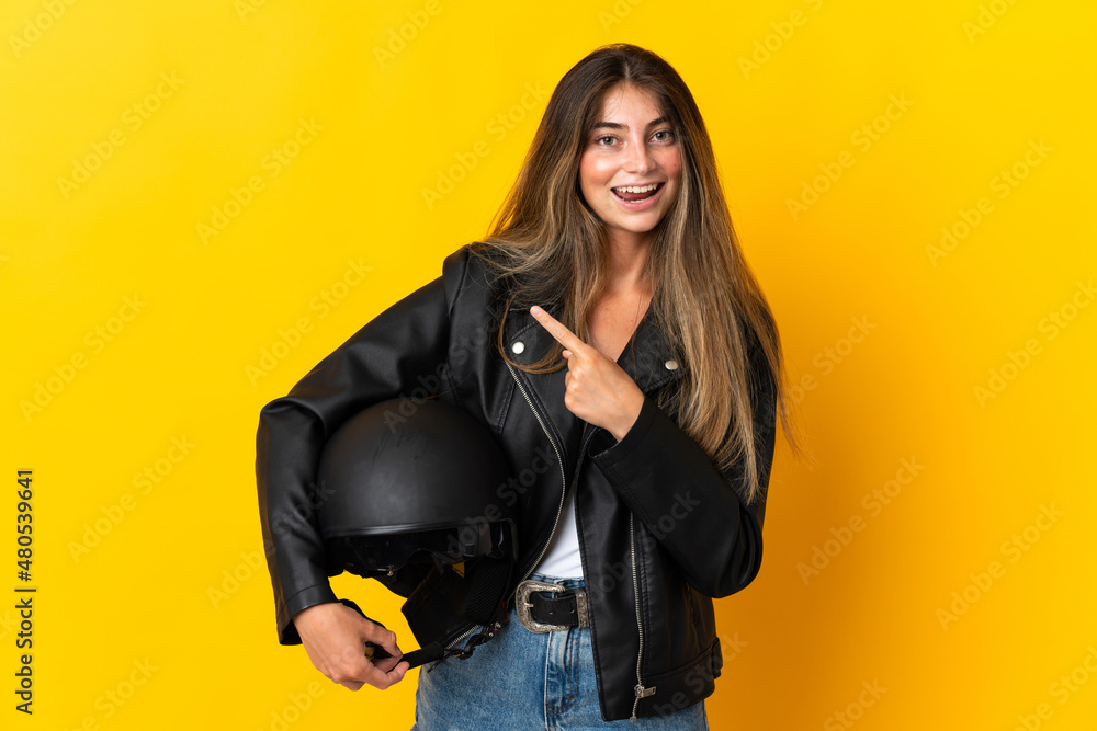 Woman holding a motorcycle helmet isolated on yellow background pointing to the side to present a product Woman holding a motorcycle helmet isolated on yellow background pointing to the side to present a product