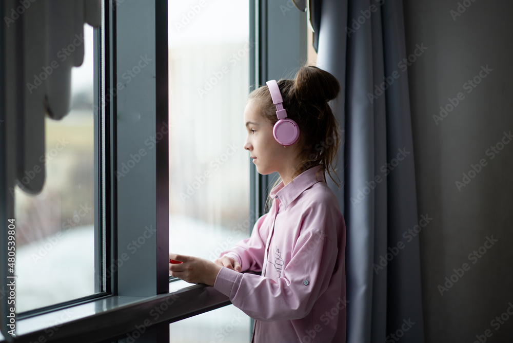 A cute teenage girl in a pink shirt and headphones looks out the window and dreams.