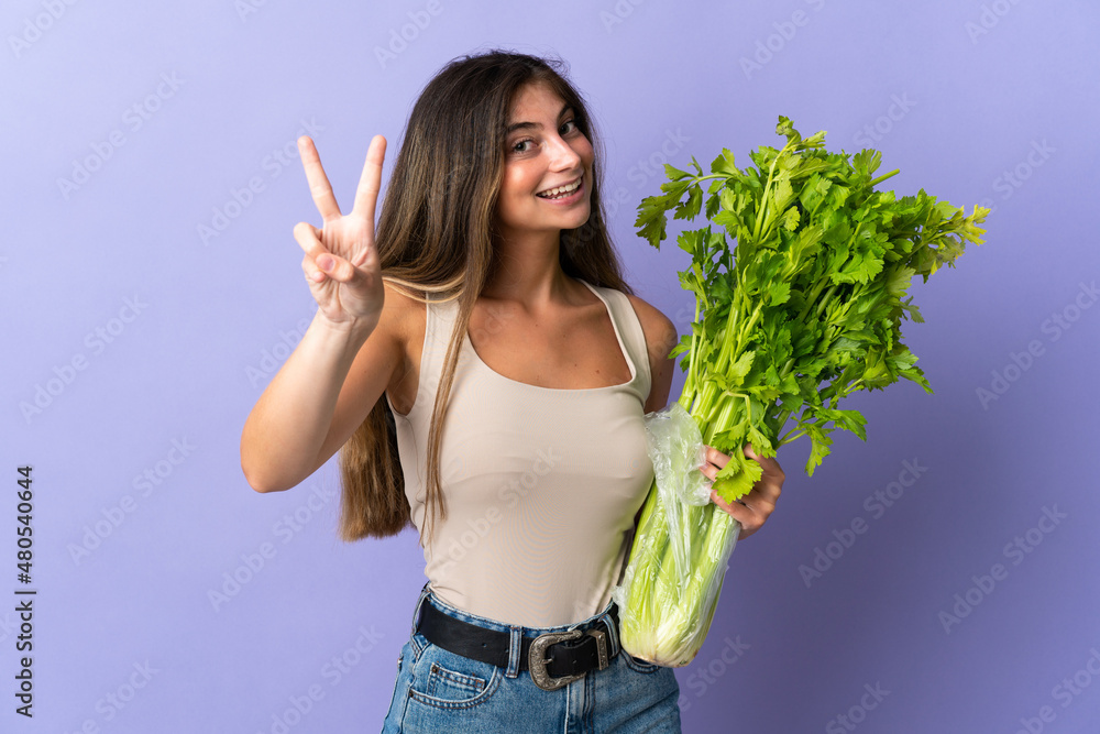 Young woman holding a celery isolated on purple background smiling and showing victory sign Young woman holding a celery isolated on purple background smiling and showing victory sign