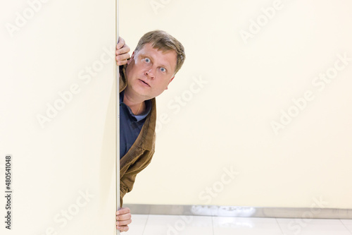 A frightened man with big eyes looks out from behind the wall.