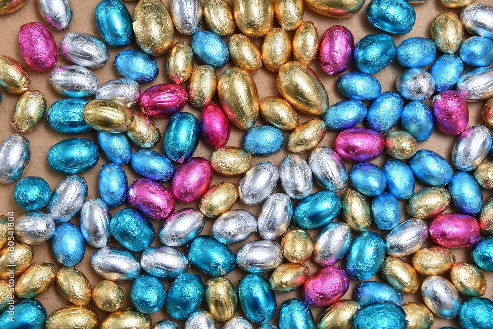 Large & small pink, blue, silver and gold foil wrapped chocolate easter eggs, against a pale wood background. Large & small pink, blue, silver and gold foil wrapped chocolate easter eggs, against a pale wood background.