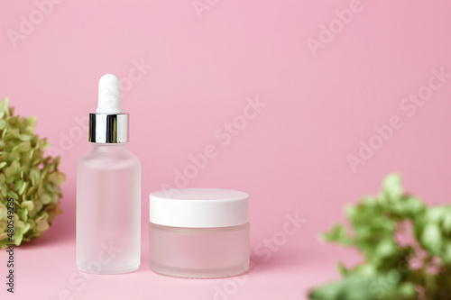 Two white glass cosmetic jars for cream and serum on a pink background with green foliage. Cosmetology and beauty. Self care. Model for cosmetics.