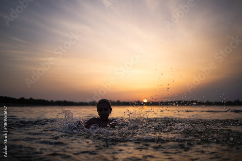 Fototapeta Naklejka Na Ścianę i Meble -  A girl plays with water in the lake and splashes it to the sides against the backdrop of a sunset